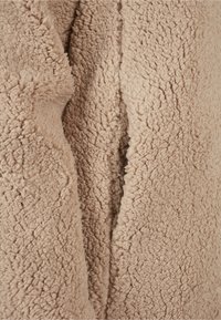 Close-up van beige zachte sherpa fleece stof met een gestructureerd, pluizig oppervlak en dichte, krullende vezels.