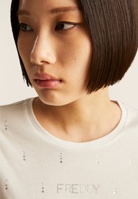 Camiseta blanca con lentejuelas y texto "FREDDY", con escote redondo y mangas cortas. Textura suave y diseño ajustado.