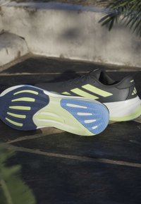 Scarpe da atleta nere con accenti gialli, tomaia testurizzata e suola multicolore con sezioni blu e verdi e modelli di battistrada distintivi.