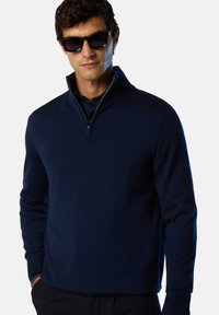 Maglione navy con zip a quarti, maniche lunghe, texture morbida e colletto alto. Indossato con occhiali da sole scuri.