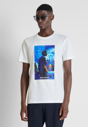 Mann trägt dunkle Sonnenbrille, weißes T-Shirt mit buntem Bild einer Person vor einem wirbelnden blauen Hintergrund und dunkle Hose.