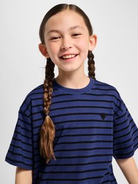 Smilende ung pige med brunt fletning hår iført en blå og sort stribet t-shirt mod en ensartet lys baggrund.