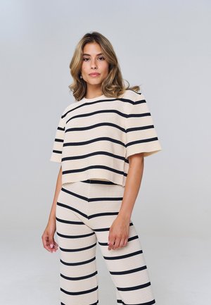 Donna con capelli biondi fino alle spalle che indossa una maglia a maniche corte beige e nera a righe orizzontali e pantaloni larghi abbinati, in piedi davanti a uno sfondo grigio.