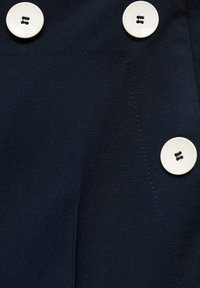Detail látky v námořnické modři, která zobrazuje plochou kapsu a dva velké, kulaté, smetanové knoflíky se čtyřmi dírkami, ukazující hladký povrch a jednoduchý design.