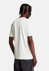 Camiseta blanca de manga corta con cuello redondo, fabricada en algodón, de textura suave y diseño minimalista. Costuras visibles en los hombros y los lados.
