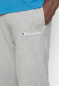 Pelēki kokvilnas sporta bikses ar izšūtu "Champion" logotipu baltā un sarkanā krāsā kreisajā augšstilbā. Iekļauj sānu kabatas un ir brīva piegriezuma.