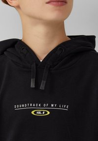 Zwarte hoodie met een trekkoord capuchon. Witte tekst met "SOUNDTRACK OF MY LIFE" en "VOL. 8" in het geel, tegen een effen achtergrond.