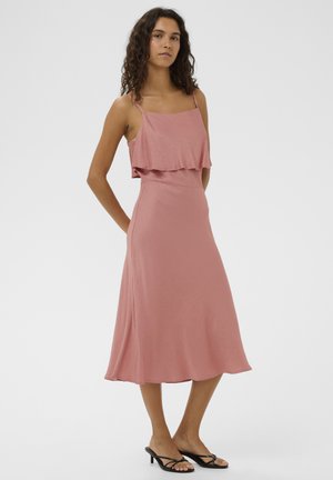 Kobieta o kręconych włosach w bezrękawowej, pudroworóżowej sukience midi i czarnych sandałach na obcasie, stojąca z rękami za plecami.