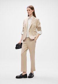 Béžový sak s klopami a zlatým gombíkom, kombinovaný so zodpovedajúcimi nohavicami a bielou prúžkovanou košeľou. Čierne kožené mokasíny a kabelka.