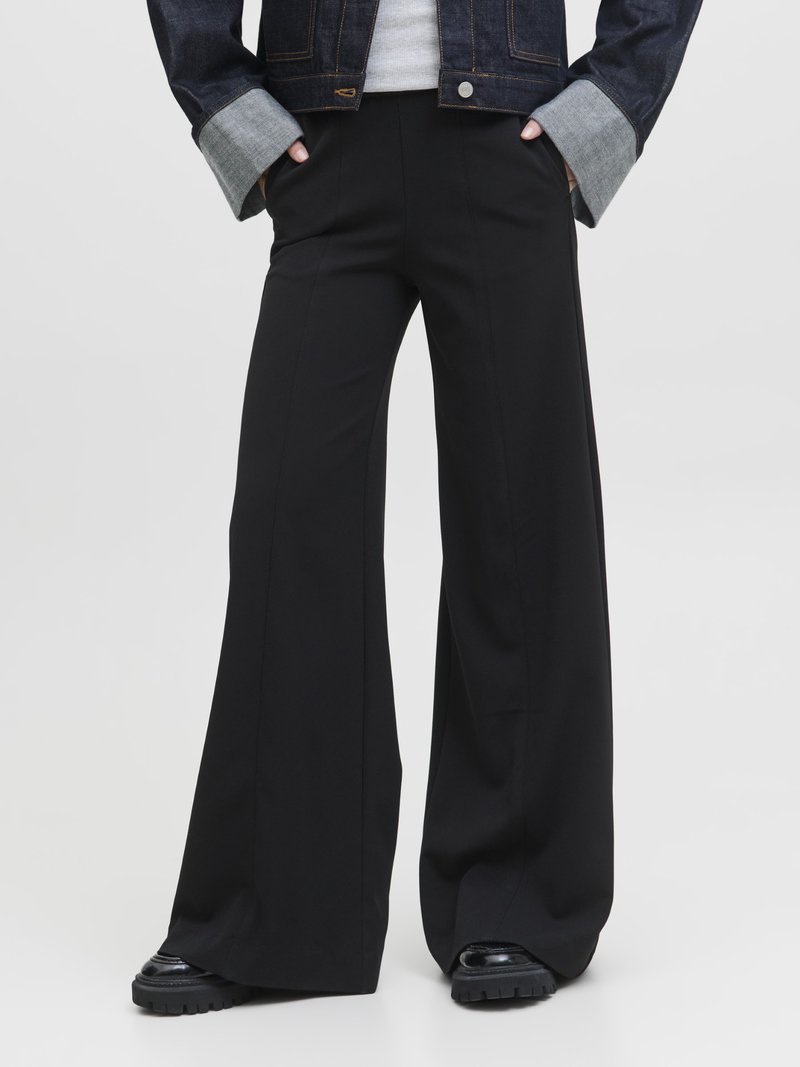 Άτομο που φοράει μαύρο φαρδύ παντελόνι, μαύρα χοντρά παπούτσια, γκρι πουκάμισο και σκουρόχρωμο τζιν μπουφάν με τα μανίκια γυρισμένα και τα χέρια στις τσέπες.