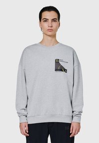 Osoba ubrana w jasnoszarą bluzę z nadrukiem na piersi oraz czarne spodnie, stojąca na tle jednolitego tła.