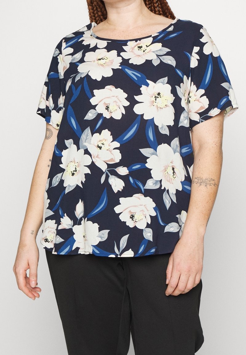 Femme portant un haut bleu marine à manches courtes avec de grands motifs floraux blancs et rose pâle et un pantalon noir, debout devant un fond uni.