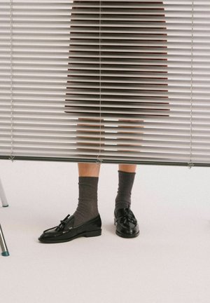 Benedenbenen van een persoon zichtbaar onder halfgesloten witte jaloezieën, met grijze sokken en zwarte loafers met kwastjes op een witte vloer.