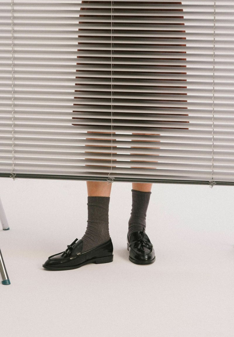 Viditelné dolní části nohou osoby pod polozavřenými bílými žaluziemi, má na sobě šedé ponožky a černé mokasíny s třásněmi na bílém podlaze.