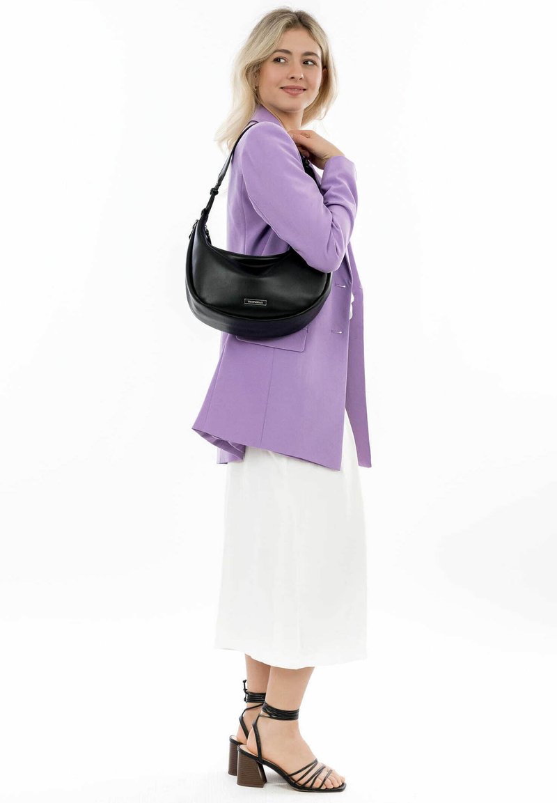 Bolso de hombro de cuero negro con forma curvada, que presenta una textura suave y un sutil detalle del logo. Llevado sobre un blazer lila claro y una falda blanca.