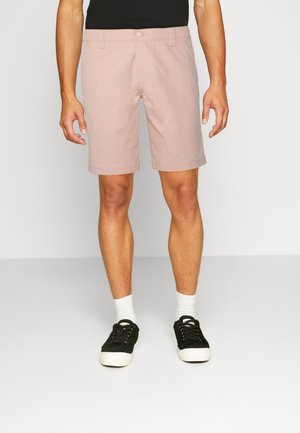 Jasnoróżowe szorty o prostym kroju, z zapięciem na guzik i bocznymi kieszeniami, w połączeniu z czarnymi trampkami i białymi skarpetkami. Gładka tekstura.