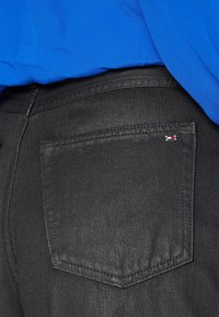 Los jeans de mezclilla negra cuentan con un bolsillo trasero que tiene una pequeña etiqueta de bandera. La tela tiene una textura suave con un ligero brillo.