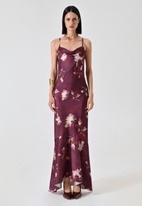 Abito lungo in satin bordeaux con motivi floreali, scollo a scialle, spalline sottili e orlo svasato. Indossato con bracciale dorato e décolleté bordeaux.