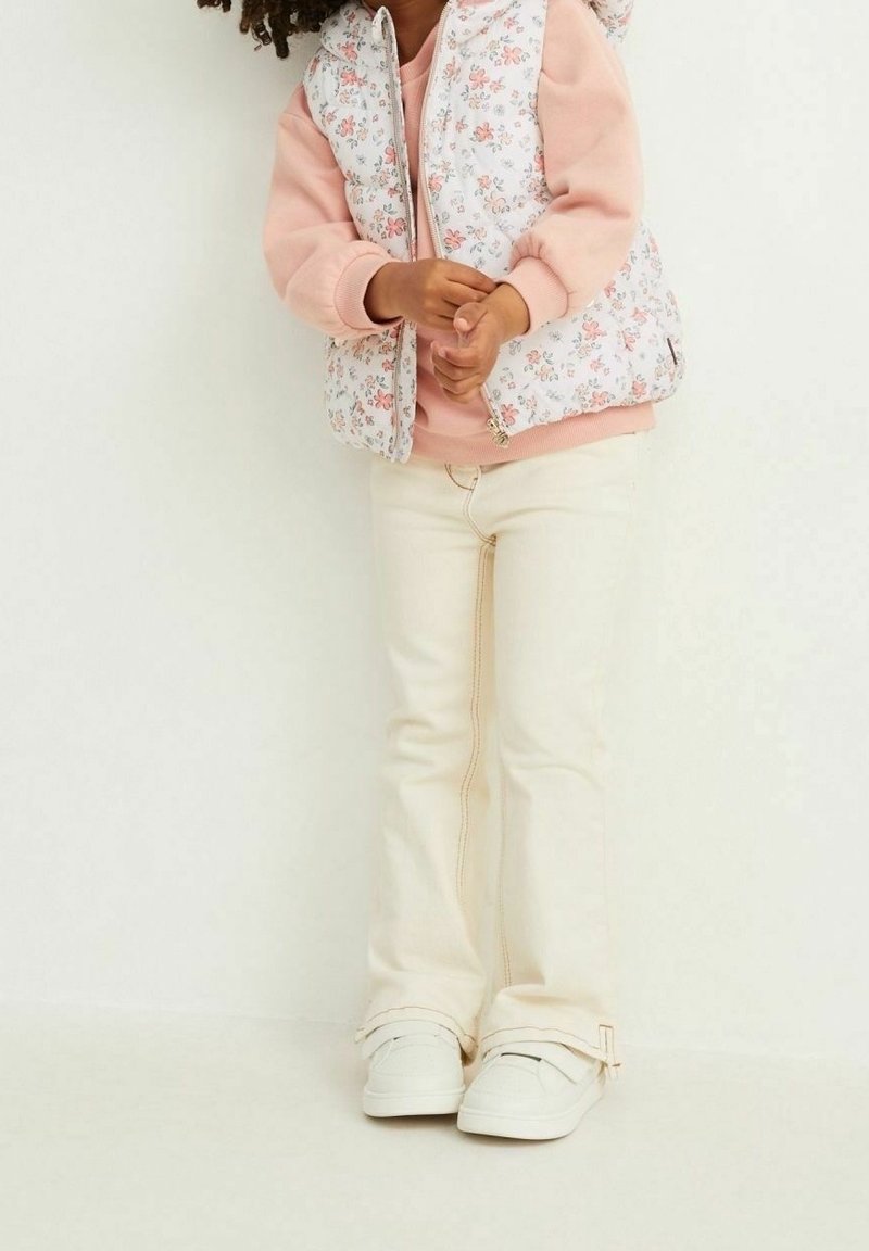 Gilet à motif floral blanc sur un sweat-shirt rose, accompagné de pantalons flare beige clair et de baskets blanches avec une bande Velcro.
