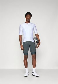 Atletinė dviračių apranga, kurioje yra balta, siaurėjančiomis juostomis megzta trumpomis rankovėmis marškinėliai ir tamsiai žali paminkštinti šortai, papildyti pilka šalmu.