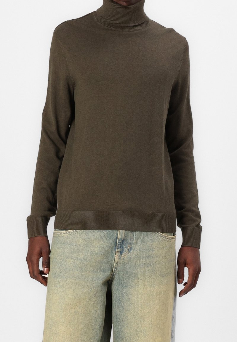 Pull à col roulé marron en matière douce, avec un col et un ourlet côtelés, porté avec un jean en denim bleu clair délavé.