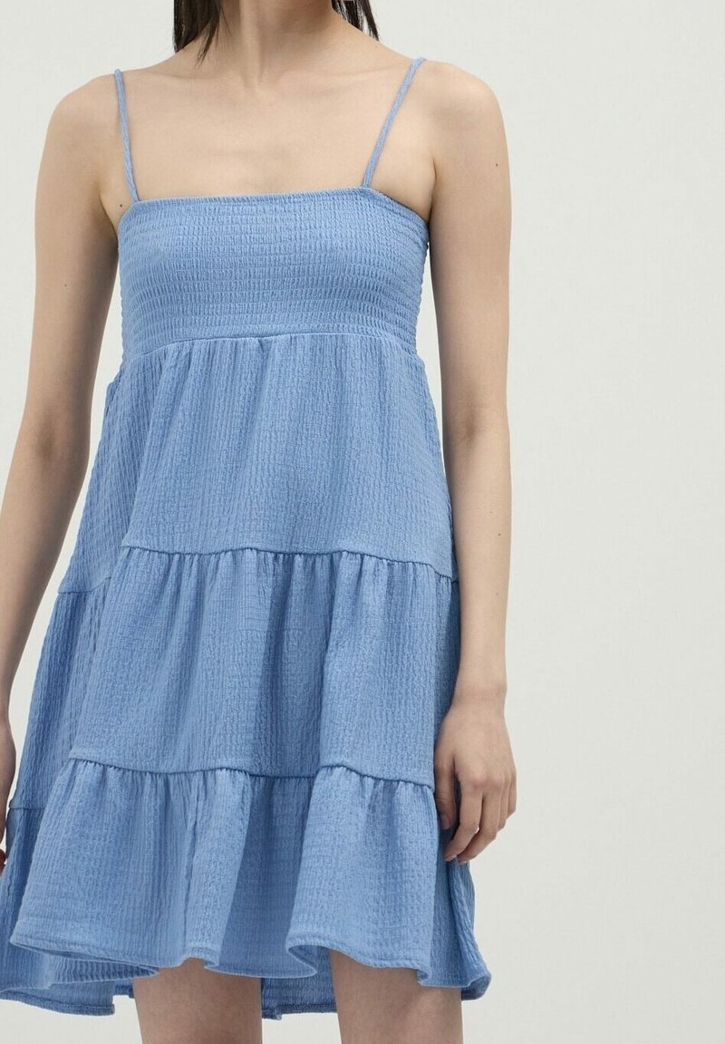 Femme portant une robe sans manches, à étages, bleu clair, avec de fines bretelles et un tissu texturé, debout devant un fond uni clair.