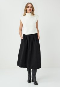Donna in piedi con le mani nelle tasche, indossa un dolcevita bianco senza maniche, una gonna midi nera e stivali neri con tacco, su uno sfondo bianco.