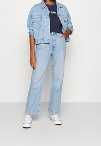 Persona vestida con una chaqueta de mezclilla azul clara y jeans, con una camiseta gráfica navy y zapatillas blancas, de pie frente a un fondo liso.