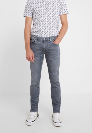 Homme portant un jean gris slim, un t-shirt blanc à motifs et des baskets blanches avec des accents noirs, debout avec une main dans la poche.