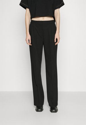 Person iført sorte bukser med høj talje og lige ben, sort cropped top og sorte sko, stående mod en ensfarvet lys baggrund.