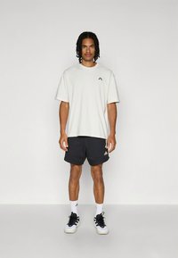 Light grey oversized t-shirt with short sleeves and small logo. Paired with dark shorts featuring white logo, and black and white athletic shoes.