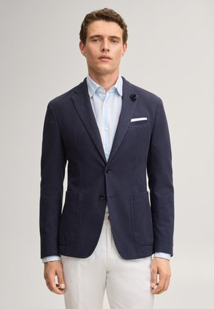 Mann trägt marineblauen Blazer über hellblau gestreiftem Hemd mit weißen Hosen, steht vor einem einfarbigen hellen Hintergrund.