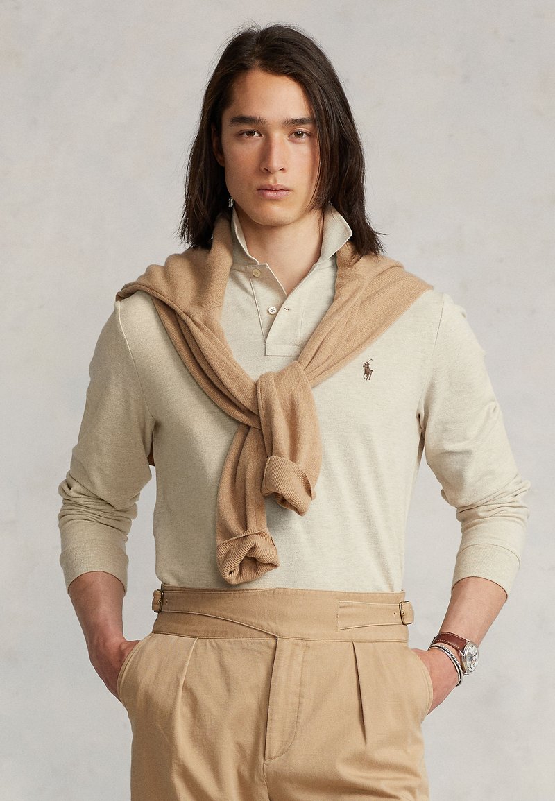 Beige collared shirt with long sleeves, featuring a small logo. A tan sweater is draped around the neck. Light brown trousers with pleats.