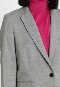 Šedý houndstooth blazer se strukturovaným střihem, se zapínáním na jeden knoflík a s klopami. Navrstvený přes jasně růžový rolák.