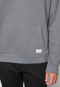 Felpa grigia con finitura testurizzata, dotata di tasca a marsupio, polsini a coste e una piccola etichetta bianca del marchio all'orlo. Pantaloni neri visibili.