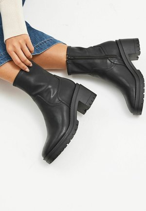 Bottines noires en cuir avec une tige flexible, fermeture éclair sur le côté, talon épais et semelle en caoutchouc texturée, présentant des finitions lisses et mates.