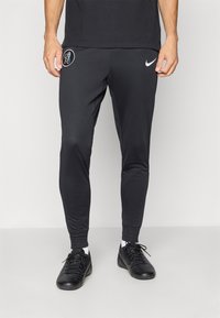 Sorte atletiske bukser lavet af glat, strækbart stof. Har et hvidt Nike-logo nær taljen og et cirkulært emblem på låret.