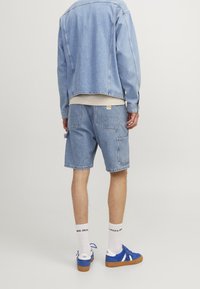 Persona che indossa una giacca di jeans azzurro chiaro e pantaloncini abbinati, calze bianche con testo nero e sneakers blu con suole color gomma, in piedi davanti a uno sfondo bianco.