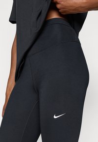 Leggings noirs extensibles avec une coupe ajustée, arborant un logo Nike blanc sur la cuisse. Texture douce et design sans coutures.