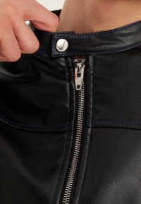 Giacca in pelle nera con cuciture blu, dotata di zip argentata e chiusura a bottone. Dettaglio ravvicinato sul colletto e sulla zip.