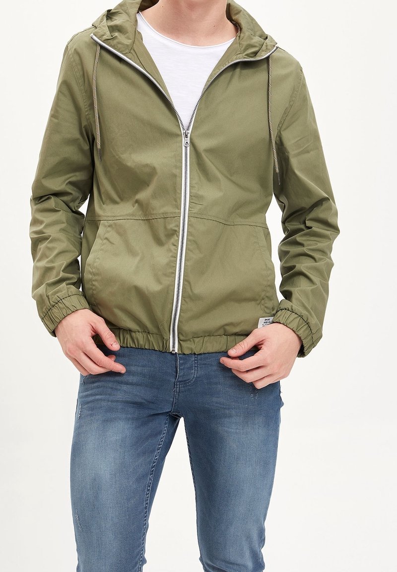 Olivgrüne leichte Jacke mit Reißverschluss, elastischen Bündchen, verstellbarem Kapuzenzug und zwei Seitentaschen, getragen über einem weißen Hemd.