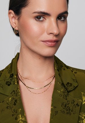 Złote naszyjniki warstwowe spoczywają na teksturowanej oliwkowo-zielonej marynarce z kwiatowymi wzorami. Marynarka ma wykładany kołnierz i subtelny połysk.