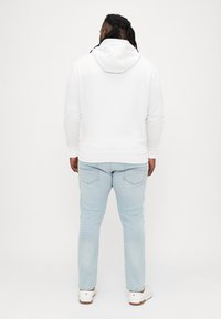 Biela mikina s kapucňou a klokaním vreckom, v kombinácii so svetlomodrými džínsami a bielymi teniskami, pohľad zo zadu.