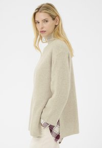 Maglione a collo alto beige a costine con una vestibilità ampia, caratterizzato da maniche lunghe e spacchi laterali. Sotto, è visibile una camicia a quadri rossa e bianca.