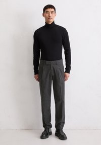Man staat in een zwart coltrui, grijze geplooide broek en zwarte veterschoenen tegen een witte muren.