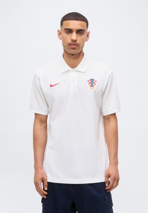 Młody mężczyzna stojący przodem, ubrany w białą koszulkę polo z czerwonym logo Nike i chorwackim herbem piłkarskim na piersi, ciemne spodnie.