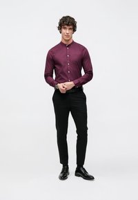 Chemise bordeaux à manches longues avec boutons et une poche, associée à un pantalon noir ajusté et des chaussures noires, portée par un mannequin aux cheveux bouclés.