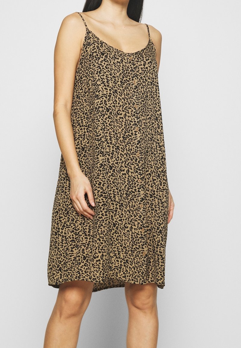 Femme portant une robe ample, sans manches, longueur genou, avec de fines bretelles et un motif léopard marron et noir.