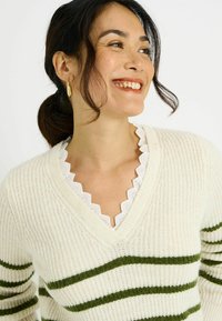 Suéter color crema con rayas horizontales verdes, hecho de tela de punto texturizada. Presenta un escote en V superpuesto sobre una blusa blanca con encaje en el borde.