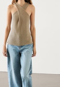 Beige knitted halter top with a crisscross design, featuring a fitted bodice and slightly flared hem, paired with light blue wide-legged jeans.